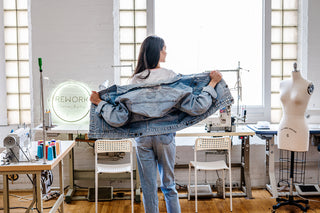 MODEL FACING BACKWARDS HOLDING OUT A JEAN JACKET WITH A SEWING STUDIO IN THE BACKGROUND